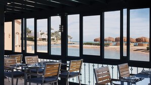A dining area with tables and chairs, large windows with a view of a beach and palm trees.