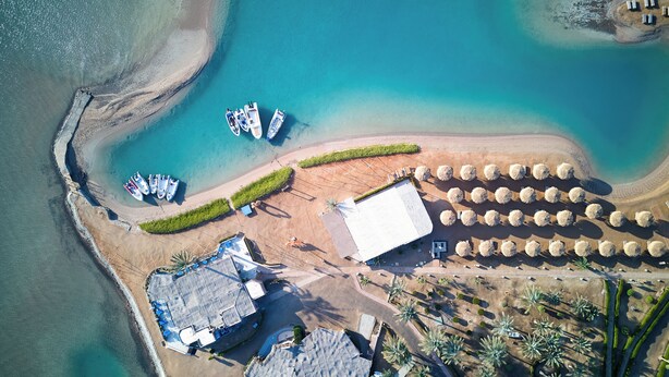 An aerial view of a beachside property with a large building, several boats docked, and a row of umbrellas.