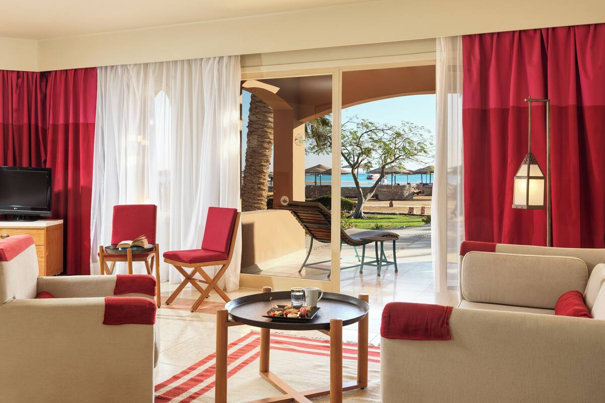 A hotel room with a TV, two red chairs, a sofa, a small table, and a view of the beach and umbrellas.