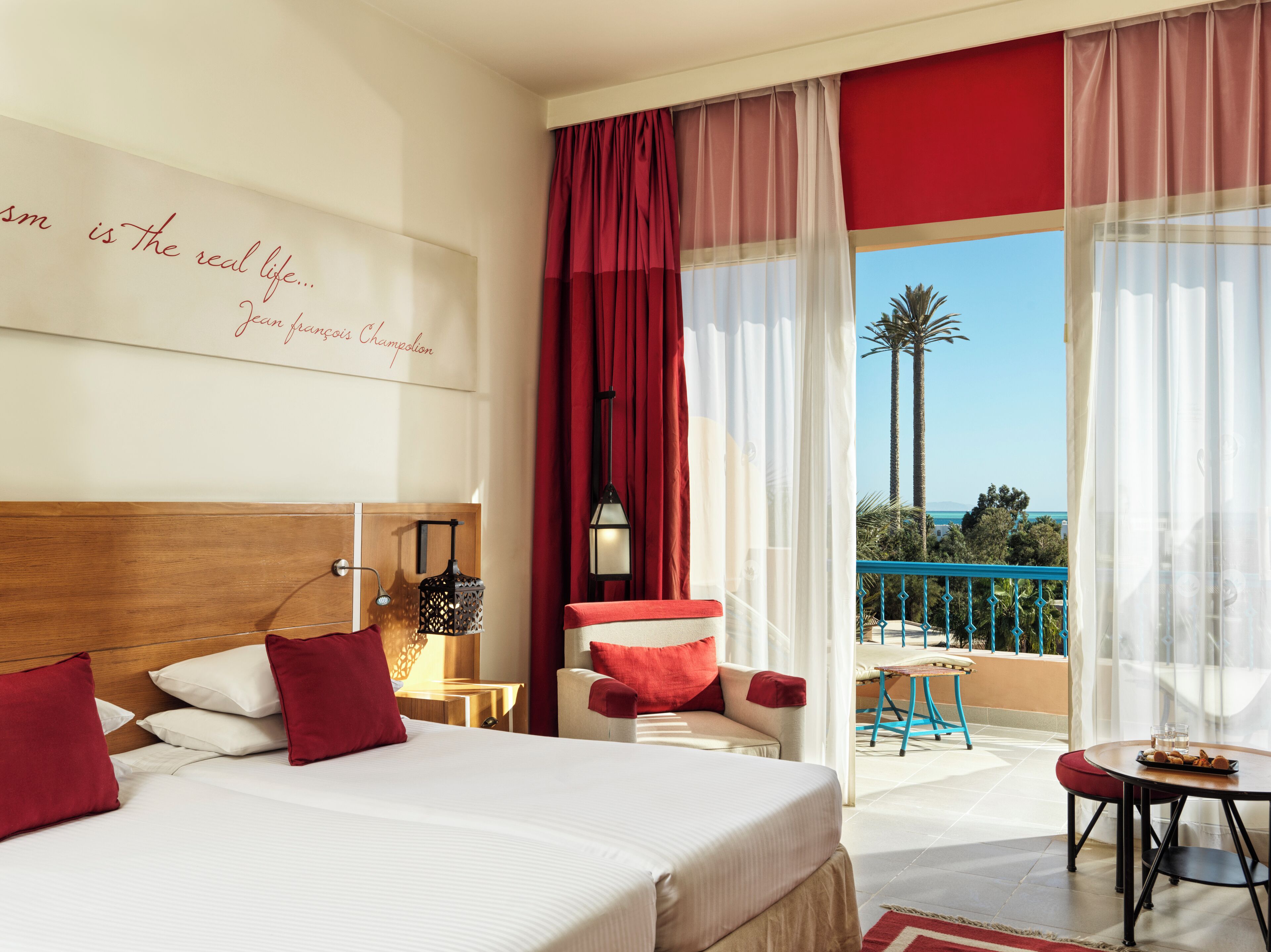Habitación de hotel con una cama grande, vista al mar, balcón con mesa y sillas, y un cartel en la pared.