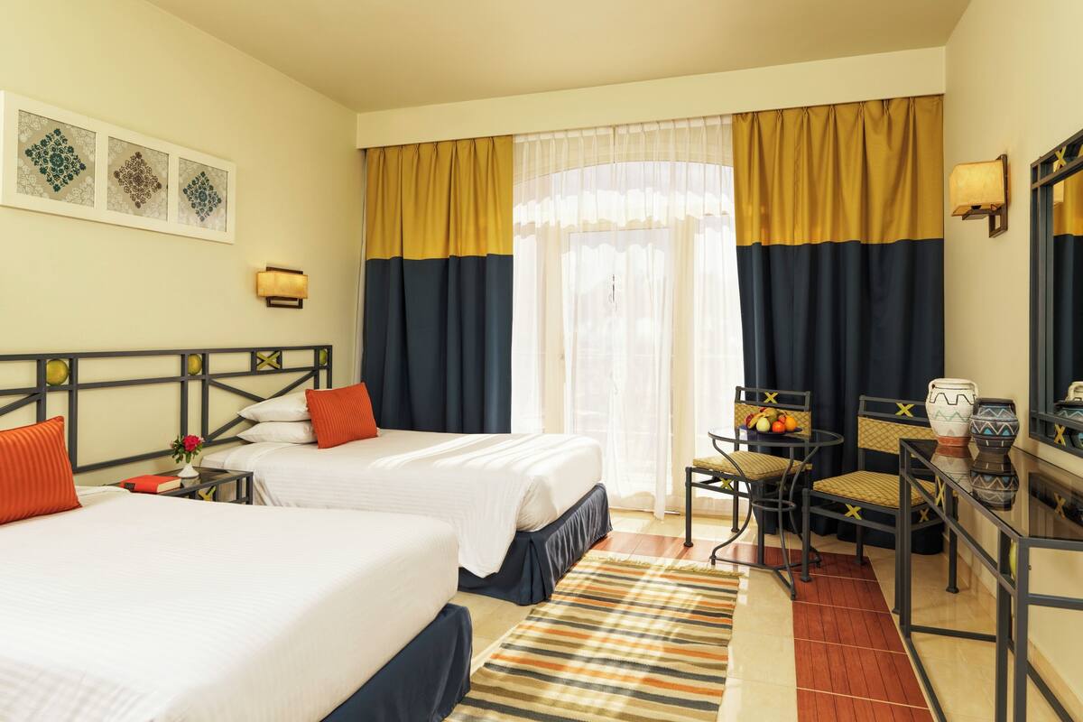 A hotel room with two beds, a desk, a chair, a small table with fruit, and a mirror.