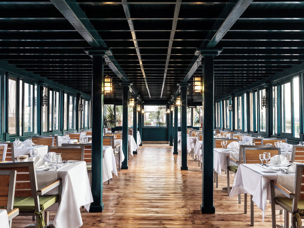 Un restaurant doté d’un long plancher en bois, de tables recouvertes de nappes blanches et de colonnes ornées de luminaires suspendus.
