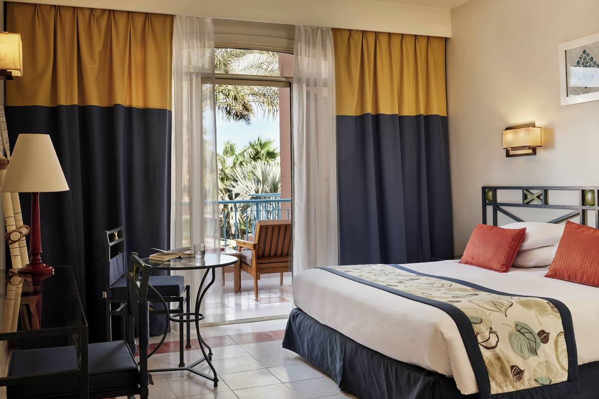 A hotel room with a bed, a desk, a chair, and a balcony with a view of palm trees.