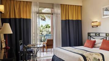 A hotel room with a bed, a desk, a chair, and a balcony with a view of palm trees.