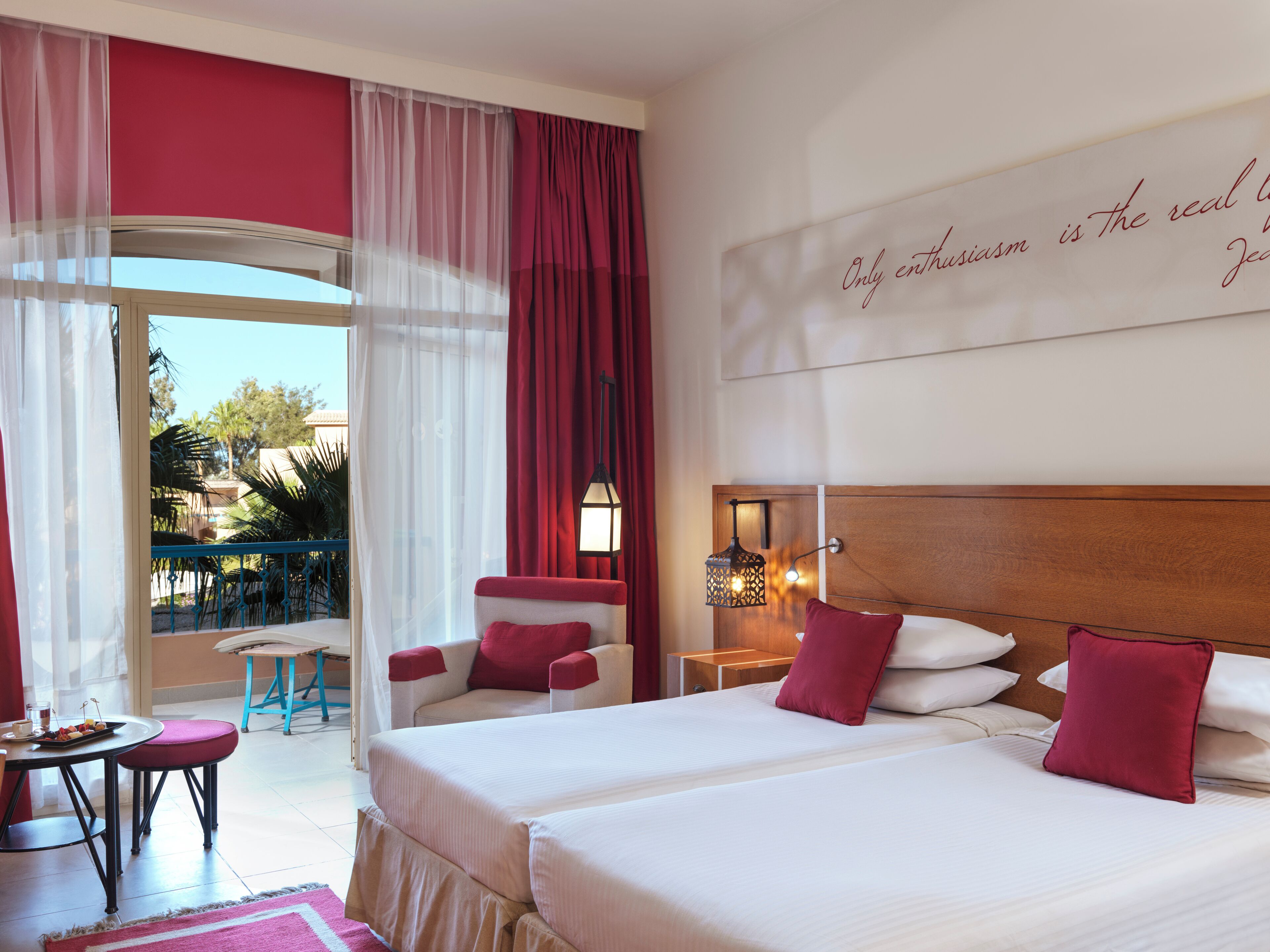 Habitación de hotel con dos camas, balcón, zona de comedor y vista a palmeras.