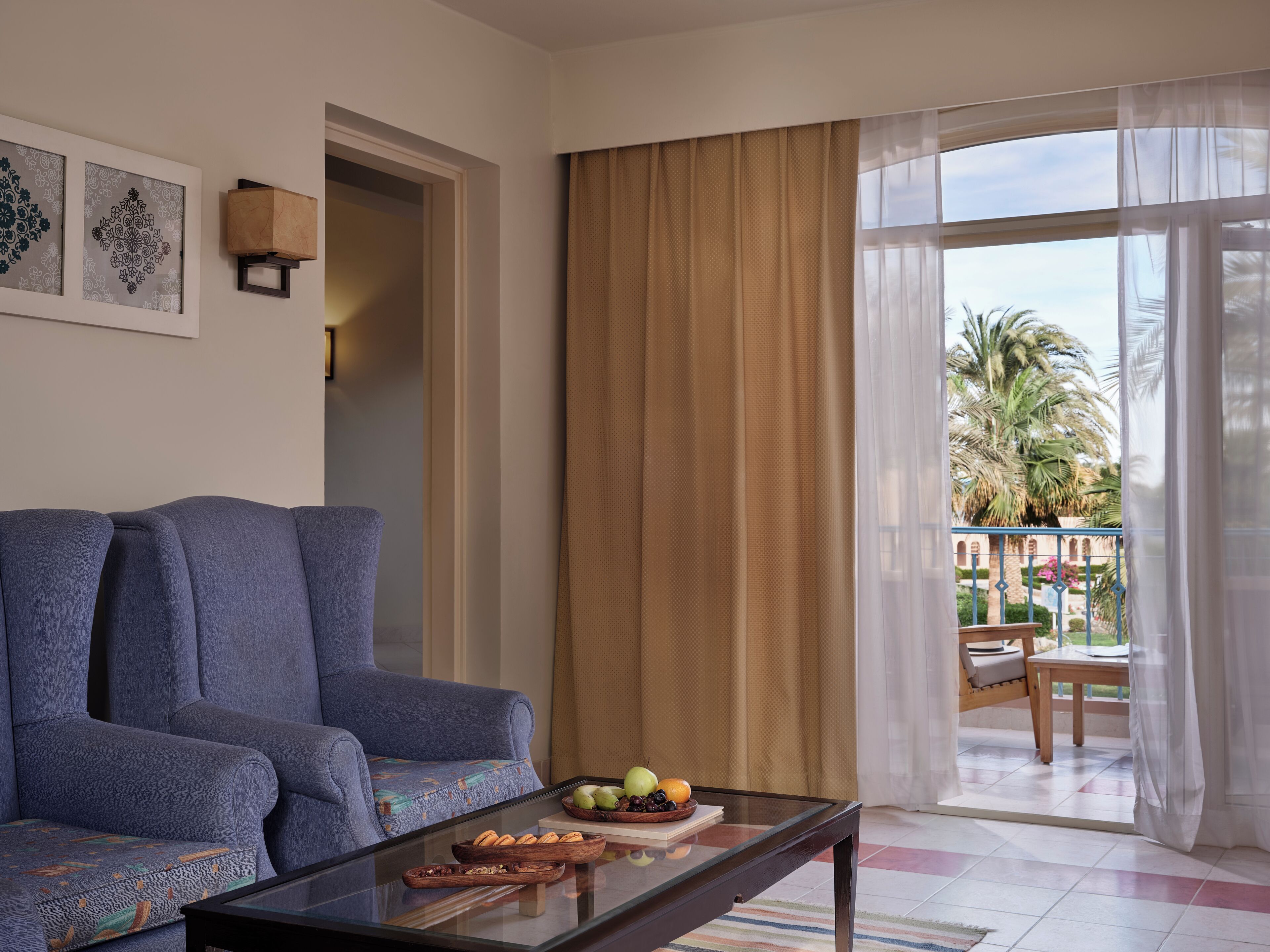 Un salón con sofá, una mesa de centro de vidrio con frutas y pan, y vistas a un balcón con palmeras.