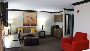 Une chambre d’hôtel avec une table à manger, un canapé, un fauteuil rouge, un coin cuisine et un tableau au mur.