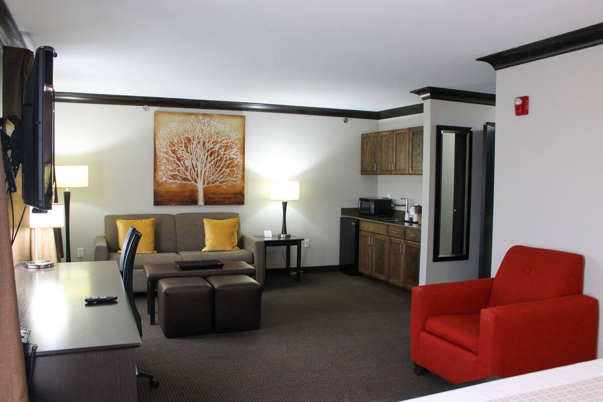 A hotel room with a dining table, a sofa, a red armchair, a kitchen area, and a painting on the wall.