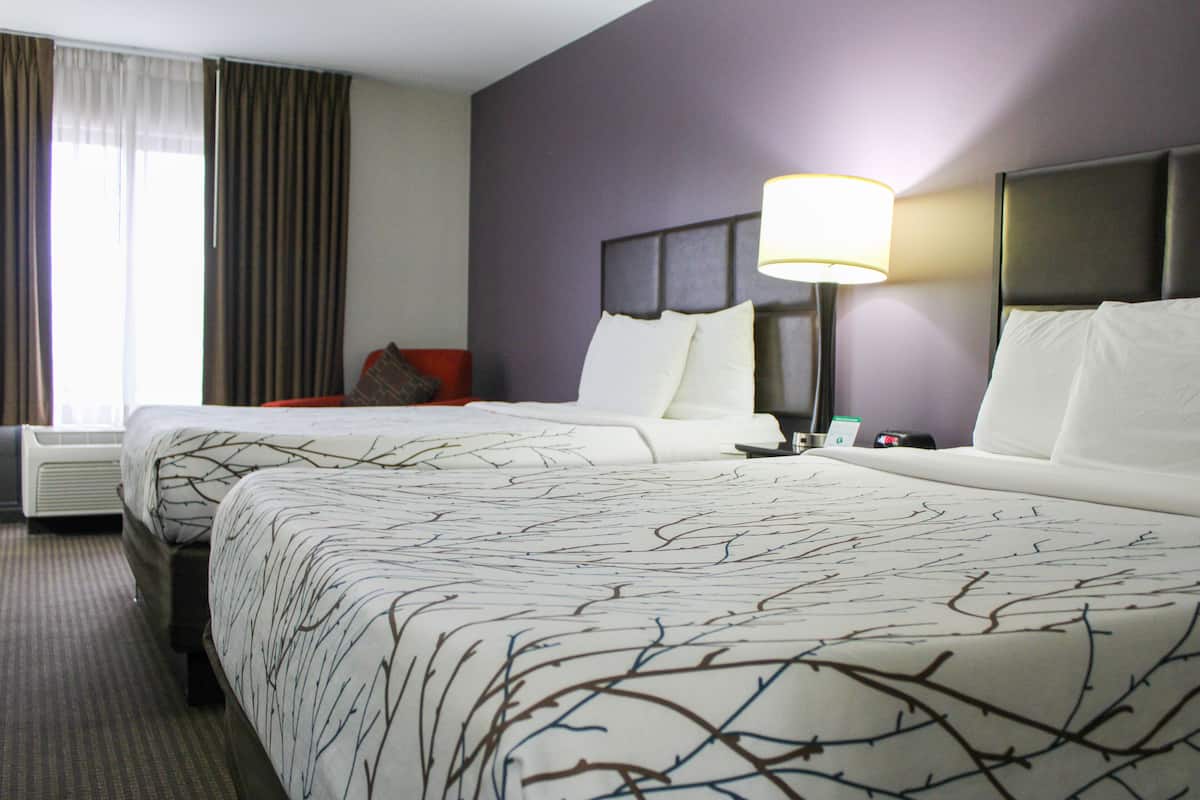A hotel room with two beds, a nightstand, a lamp, and a window with curtains.
