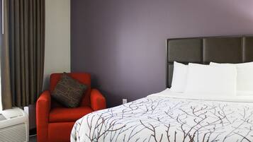 A hotel room with a large bed, a red armchair, a wall-mounted heater, and a window with curtains.