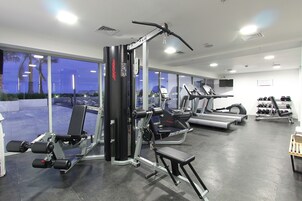 Un gimnasio con diversos equipos de ejercicio, grandes ventanales y vistas al exterior.