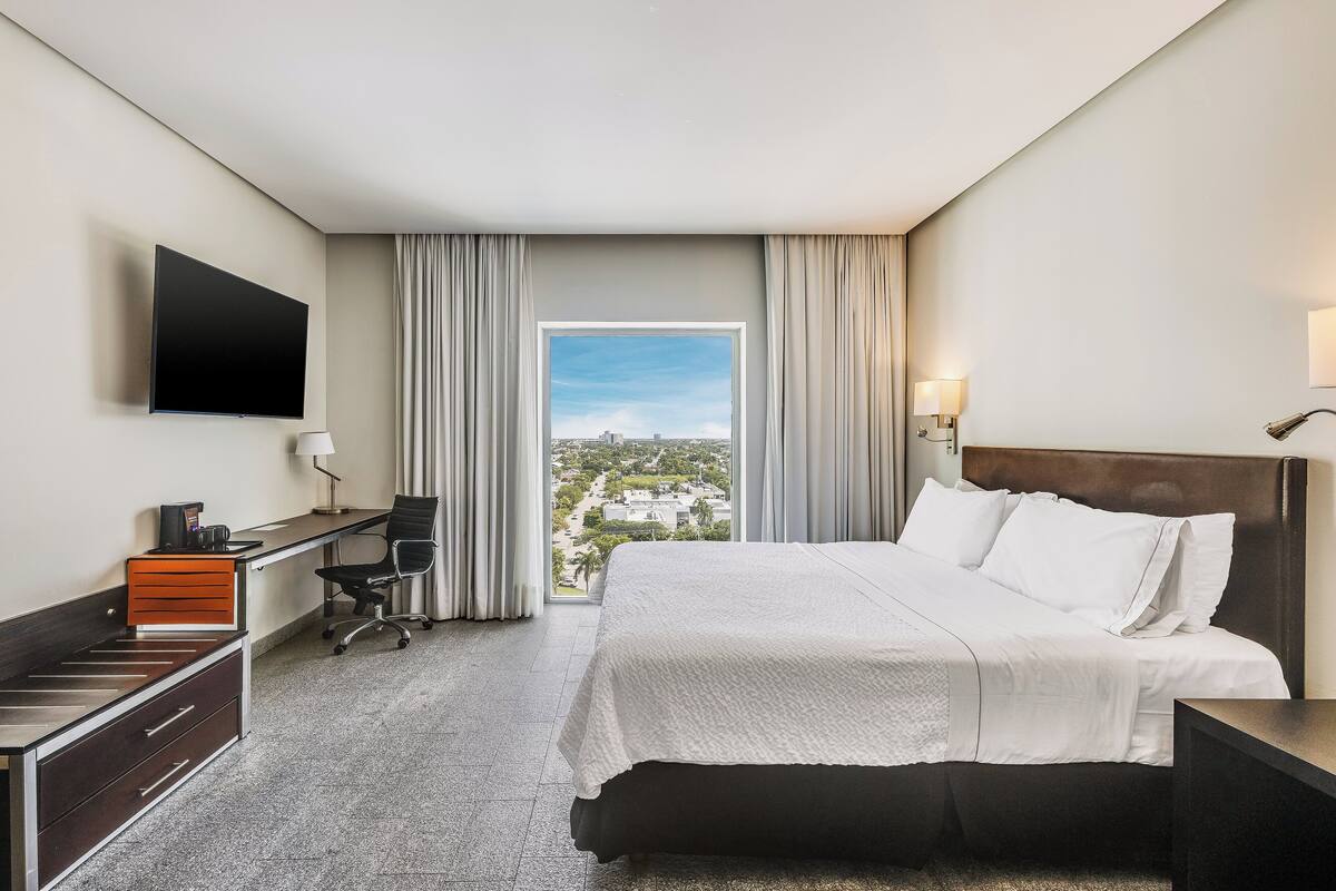 Una habitación de hotel con cama, escritorio, silla, televisor y ventana con cortinas.