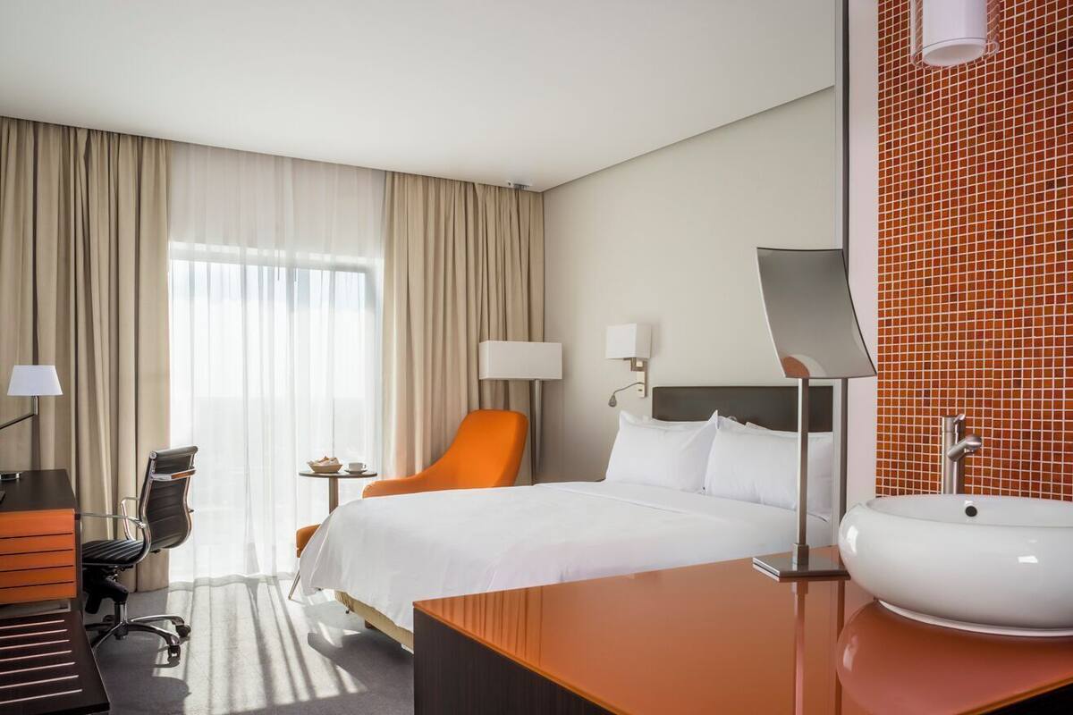 Una habitación de hotel con una cama, una silla naranja, un escritorio con lámpara, una mesita pequeña y un baño con lavabo y espejo.