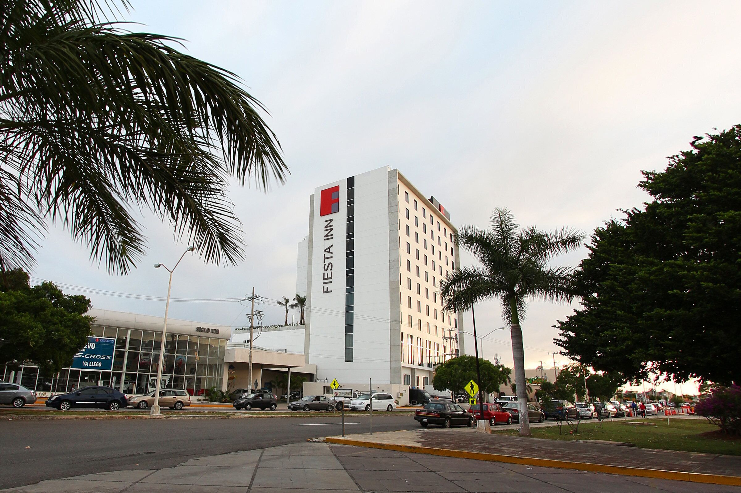 Un hotel alto con un llamativo letrero 'Fiesta', fachada de vidrio moderna y un estacionamiento con varios coches.