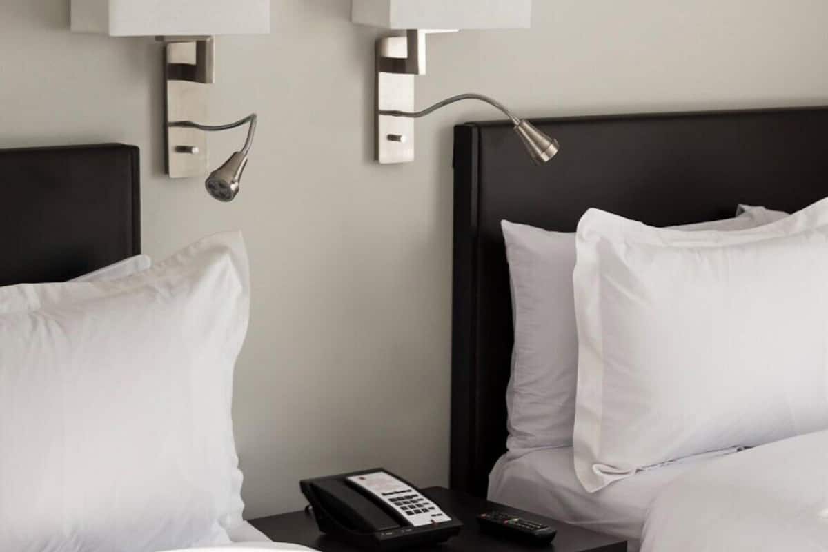 Habitación de hotel con dos camas, cada una con ropa blanca, cabeceros oscuros, una mesita de noche con teléfono y control remoto, y dos lámparas de pared.