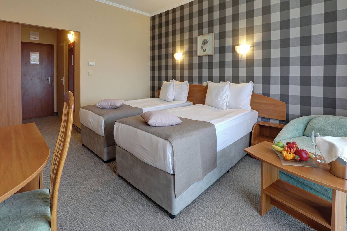 A hotel room with two beds, a wooden desk, a chair, and a small table with fruits and a glass.