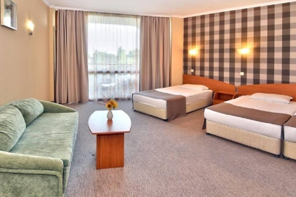 A hotel room with a green sofa, two single beds, a wooden headboard, a small table with a vase, and a view of the outdoors through large windows.