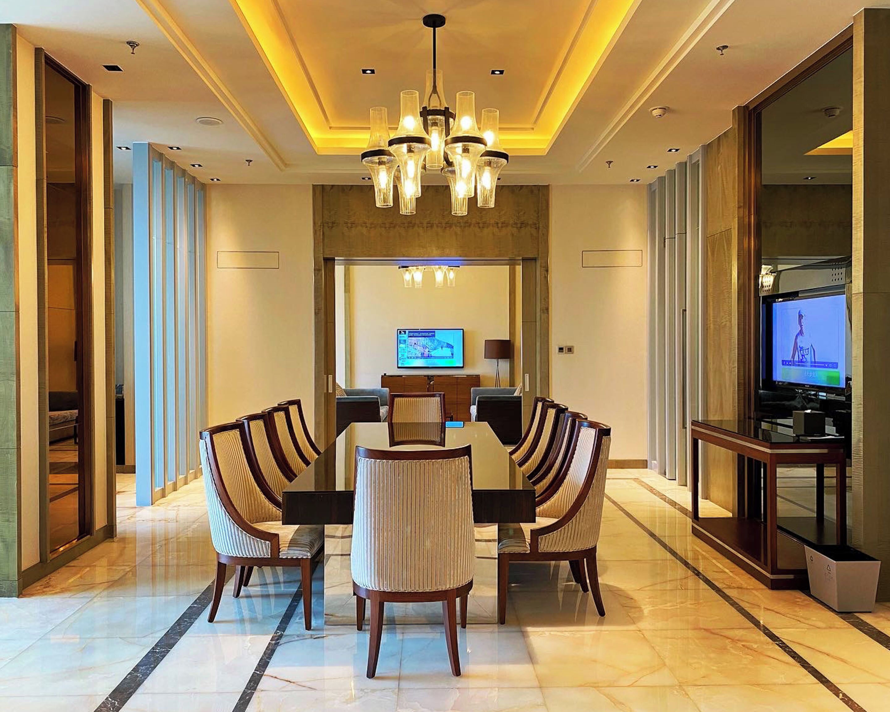 A hotel dining area with a large table, four chairs, a chandelier, a television, and a modern interior design.