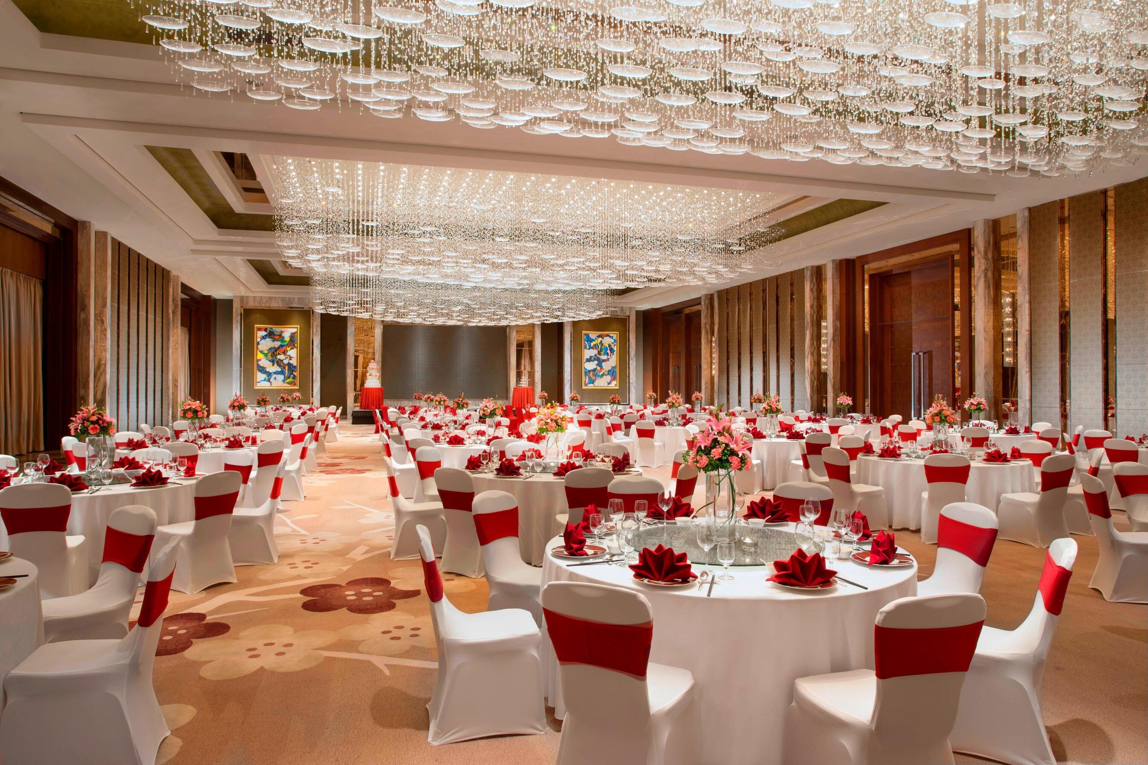 A banquet hall with round tables, white chairs with red sashes, and a large chandelier.