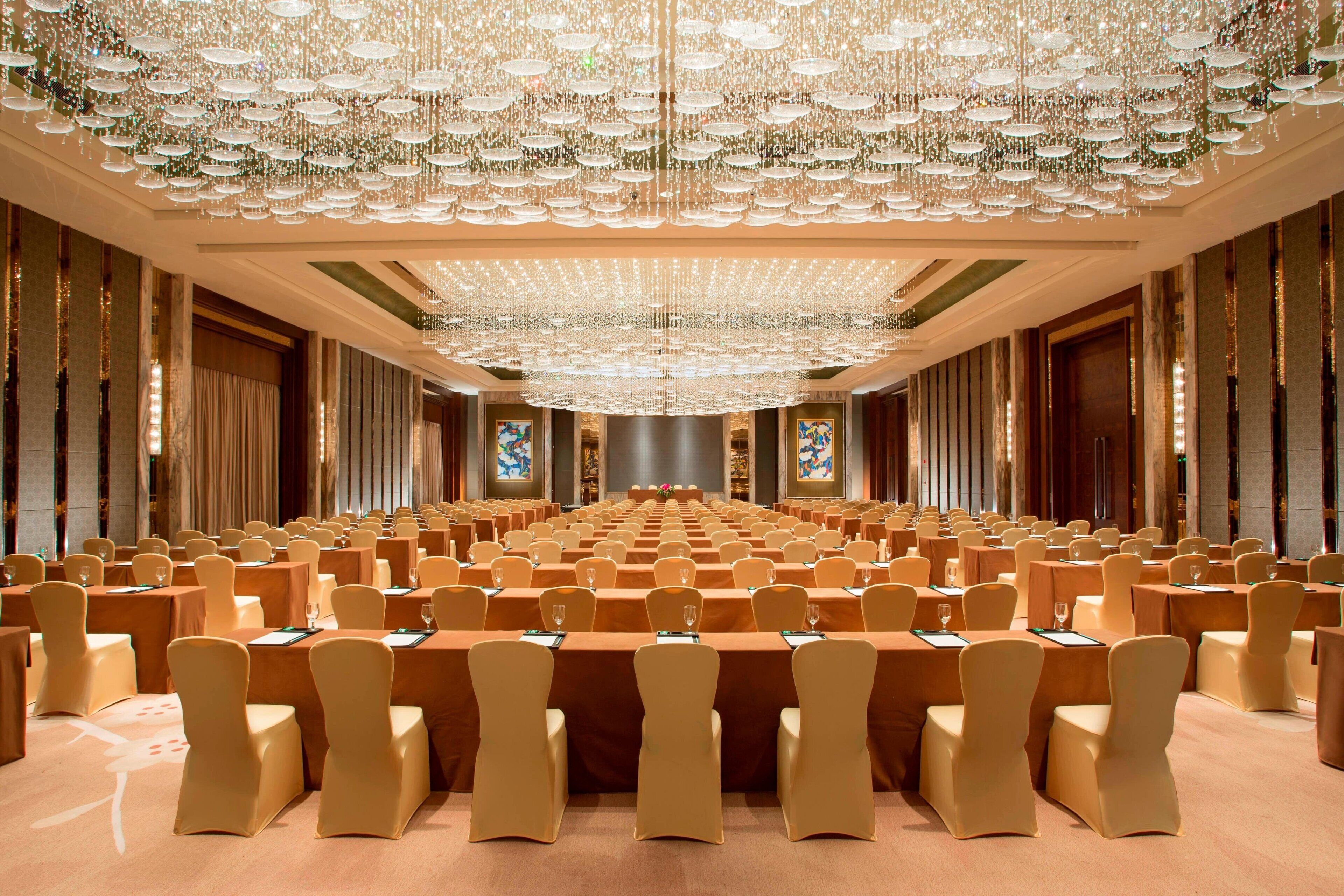 A large conference room with rows of tables and chairs, a grand chandelier, and large windows with curtains.