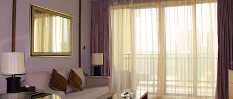 Habitación de hotel con sofá, una mesita de vidrio, lámpara y un ventanal con cortinas translúcidas.