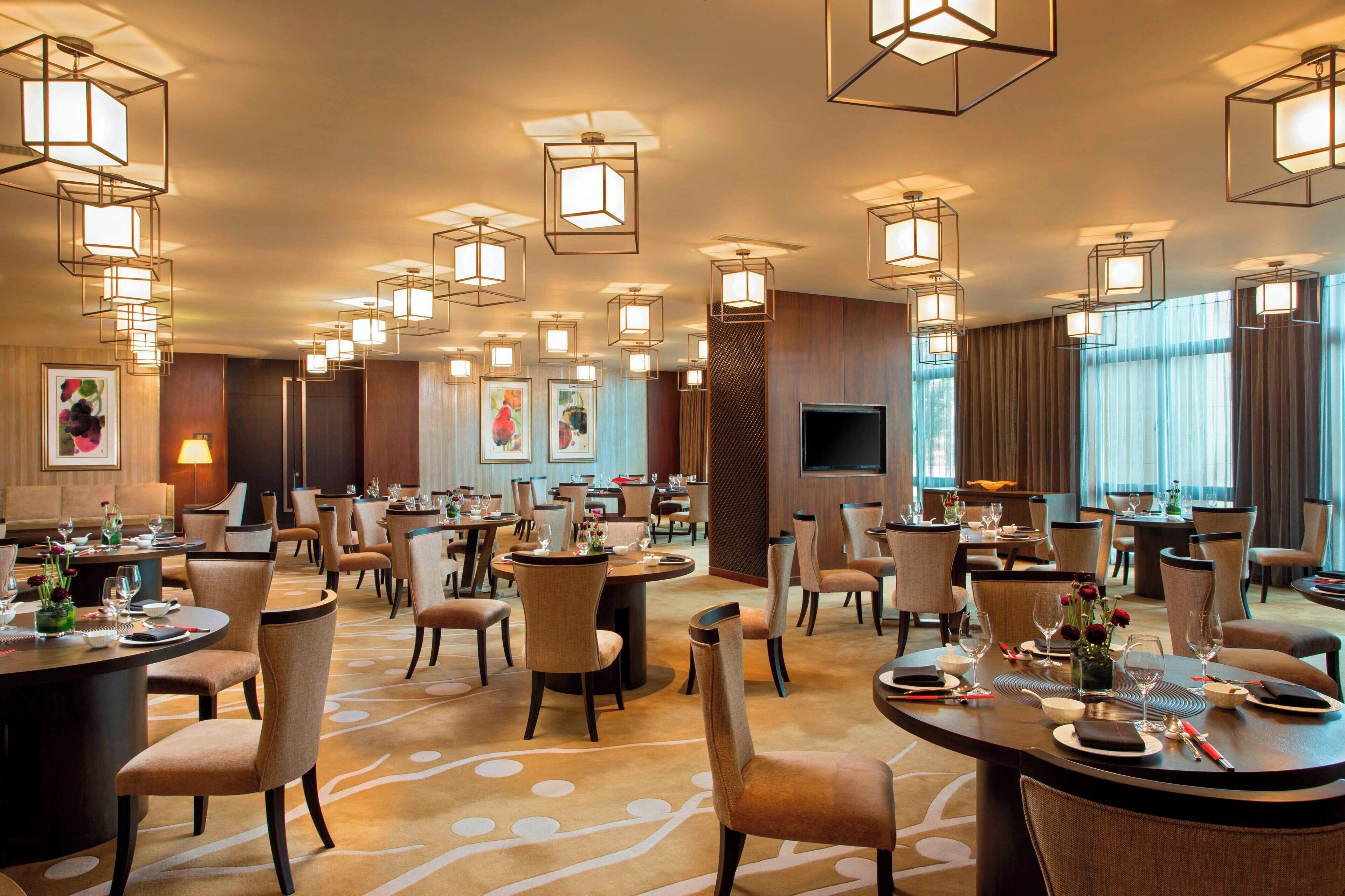 A hotel dining area with multiple round tables, chairs, and pendant lighting.