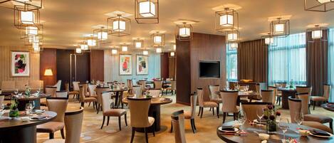 A hotel dining area with multiple round tables, chairs, and pendant lighting.