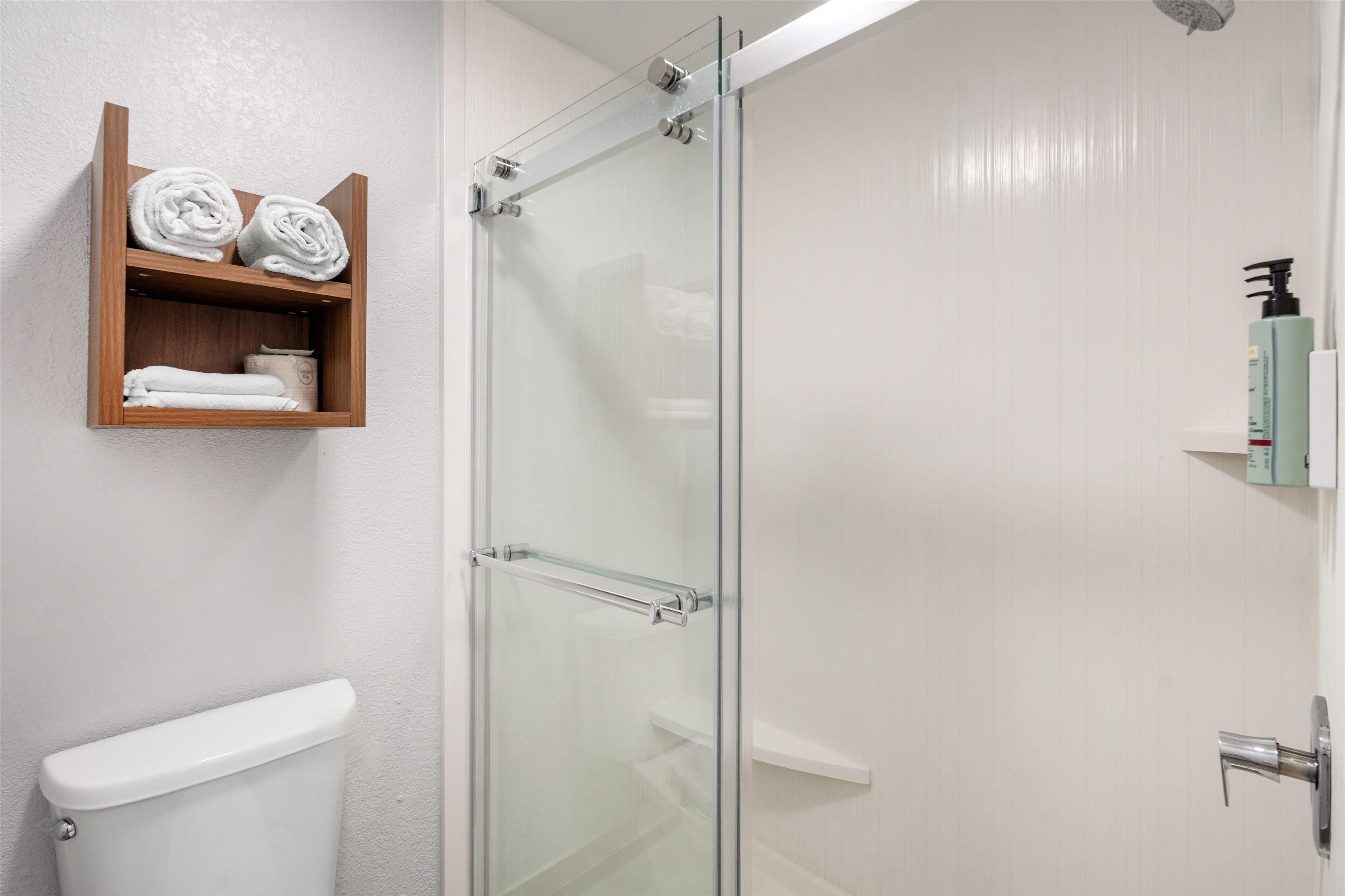 A bathroom with a glass-enclosed shower, a toilet, and a wooden shelf with towels.