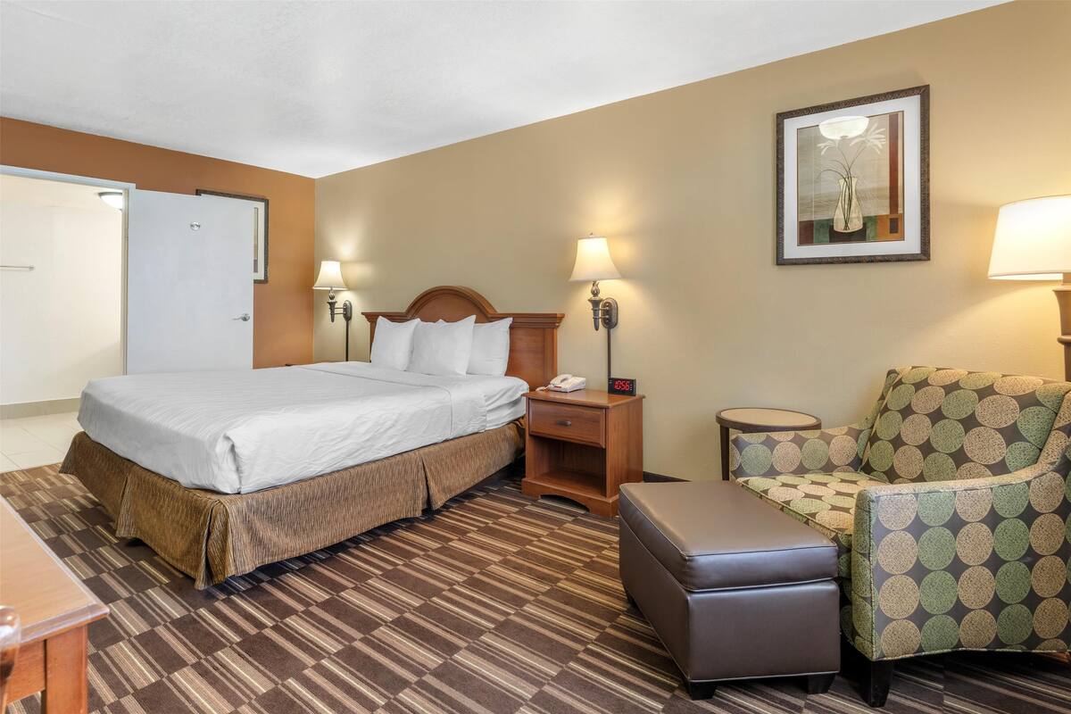 A hotel room with a bed, bedside table, lamp, chair, and a small table.