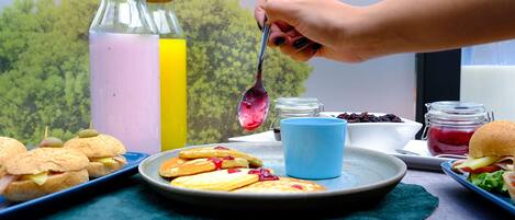 A table with food items including sandwiches, pancakes, and a burger, with a bottle of pink liquid and a blue cup in the background.
