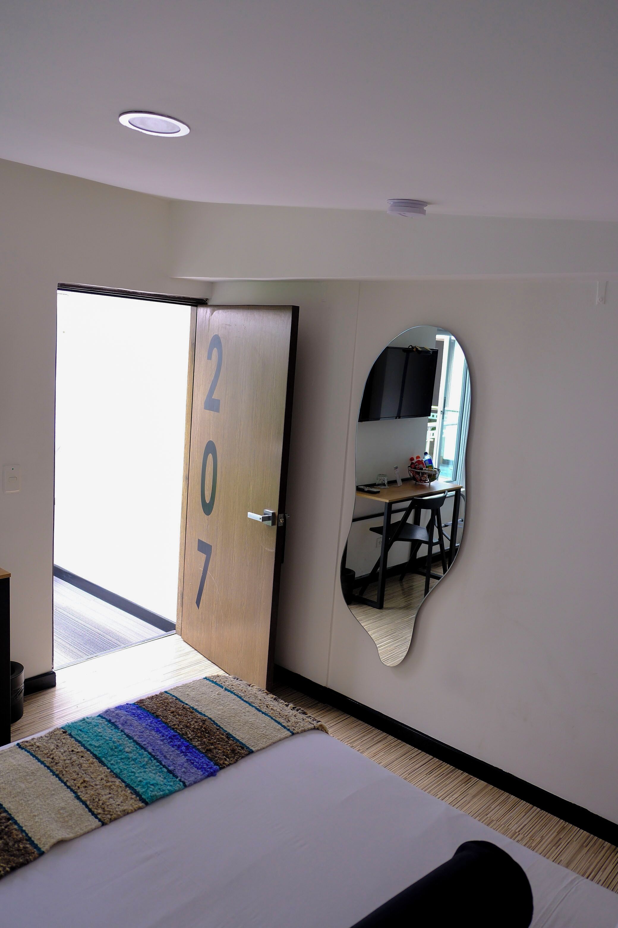 A hotel room with a bed, a mirror, a door with numbers, and a view of a dining area.