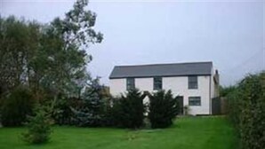 A two-story house with a white exterior and dark roof, surrounded by a well-maintained lawn and dense shrubbery. - The Woodlands (Ely)