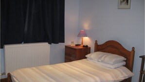 A single bed with a wooden headboard, a bedside table with a lamp, and a window with curtains. - The Woodlands (Ely)
