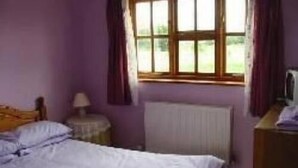 A bedroom with a bed, a nightstand, a lamp, a radiator, and a window with curtains. - The Woodlands (Ely)