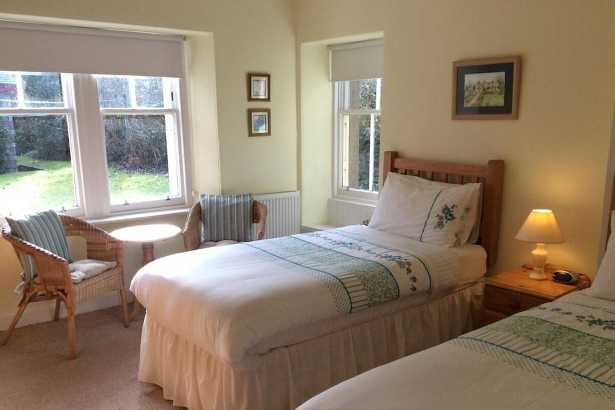 A bedroom with two beds, a chair, a bedside table with a lamp, and a window.