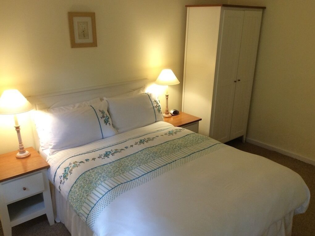 Una cama bien hecha con colcha estampada, dos lámparas de noche, una mesita de noche y un armario en una habitación con paredes lisas.