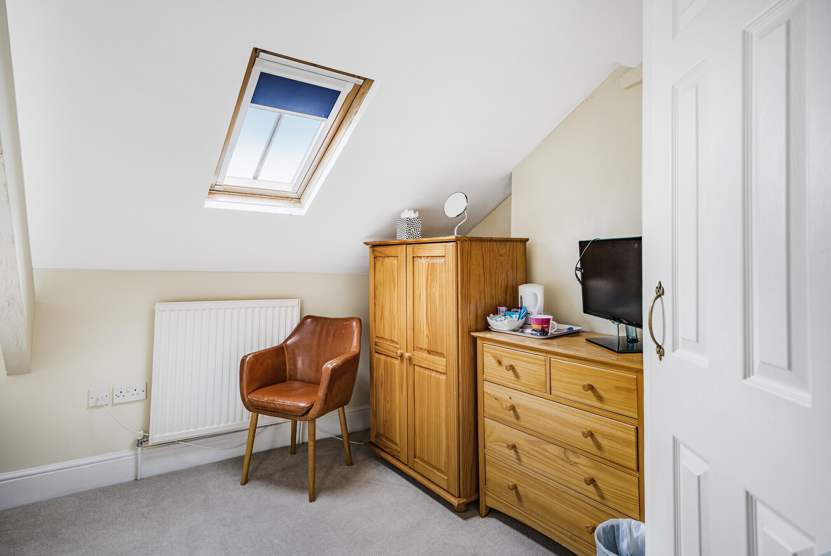 Una habitación con techo inclinado, un armario de madera, un sillón de cuero marrón, un televisor sobre una cómoda y una ventana con vistas al cielo.