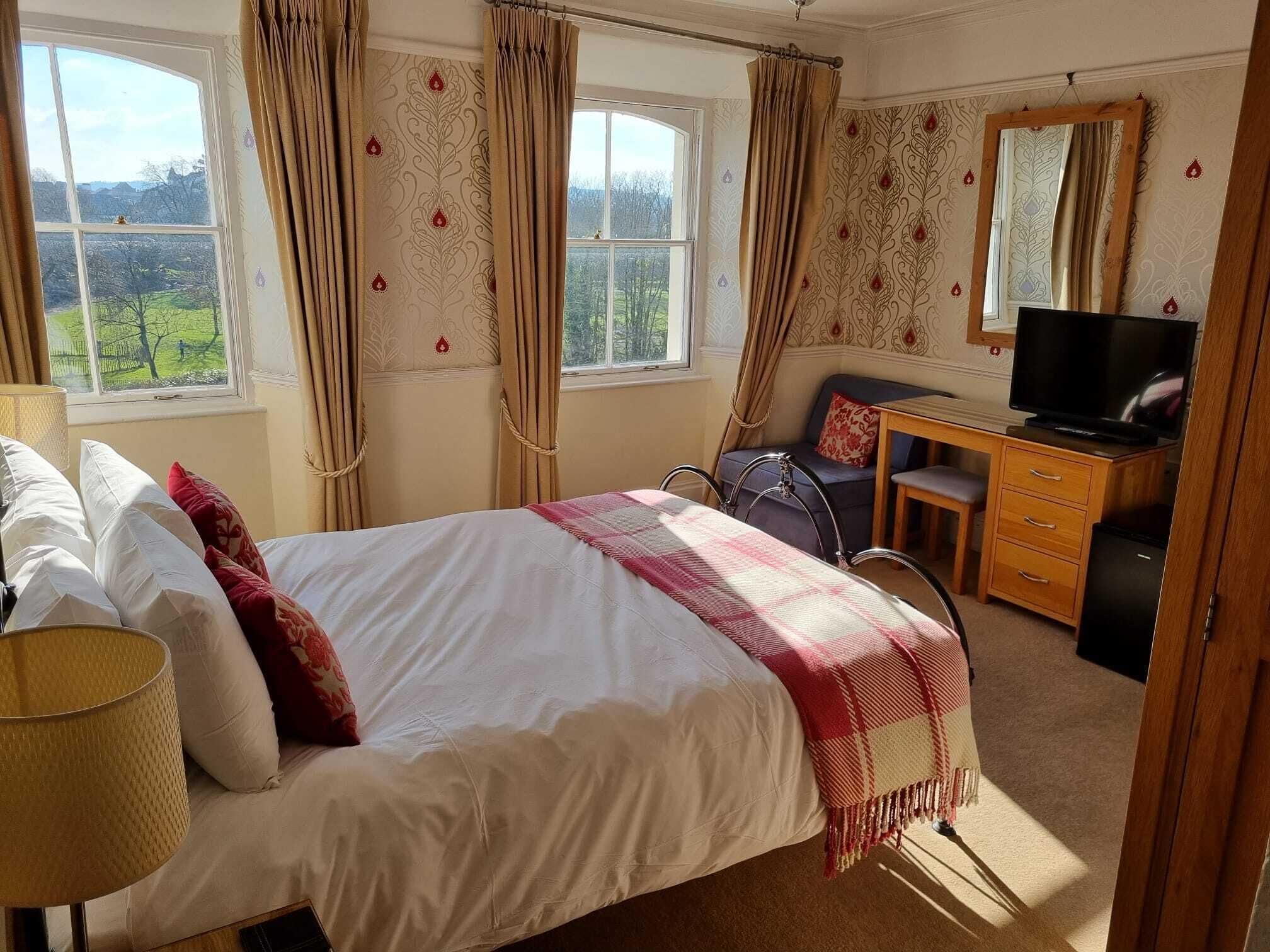 A bedroom with a bed, a desk, a chair, a television, and a view of the outdoors.
