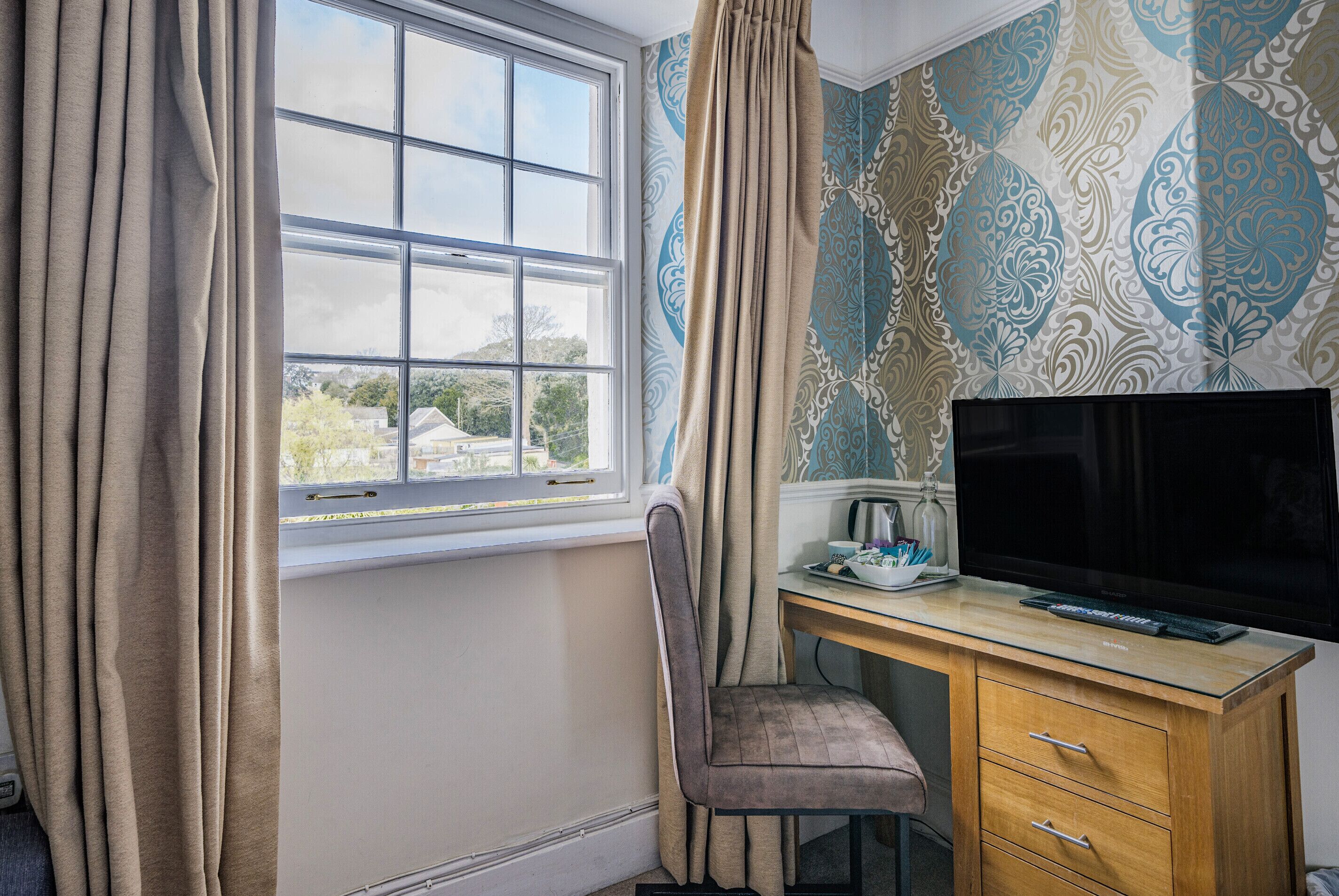 A room with a large window, a desk with a flat-screen TV, a chair, and a wooden cabinet.
