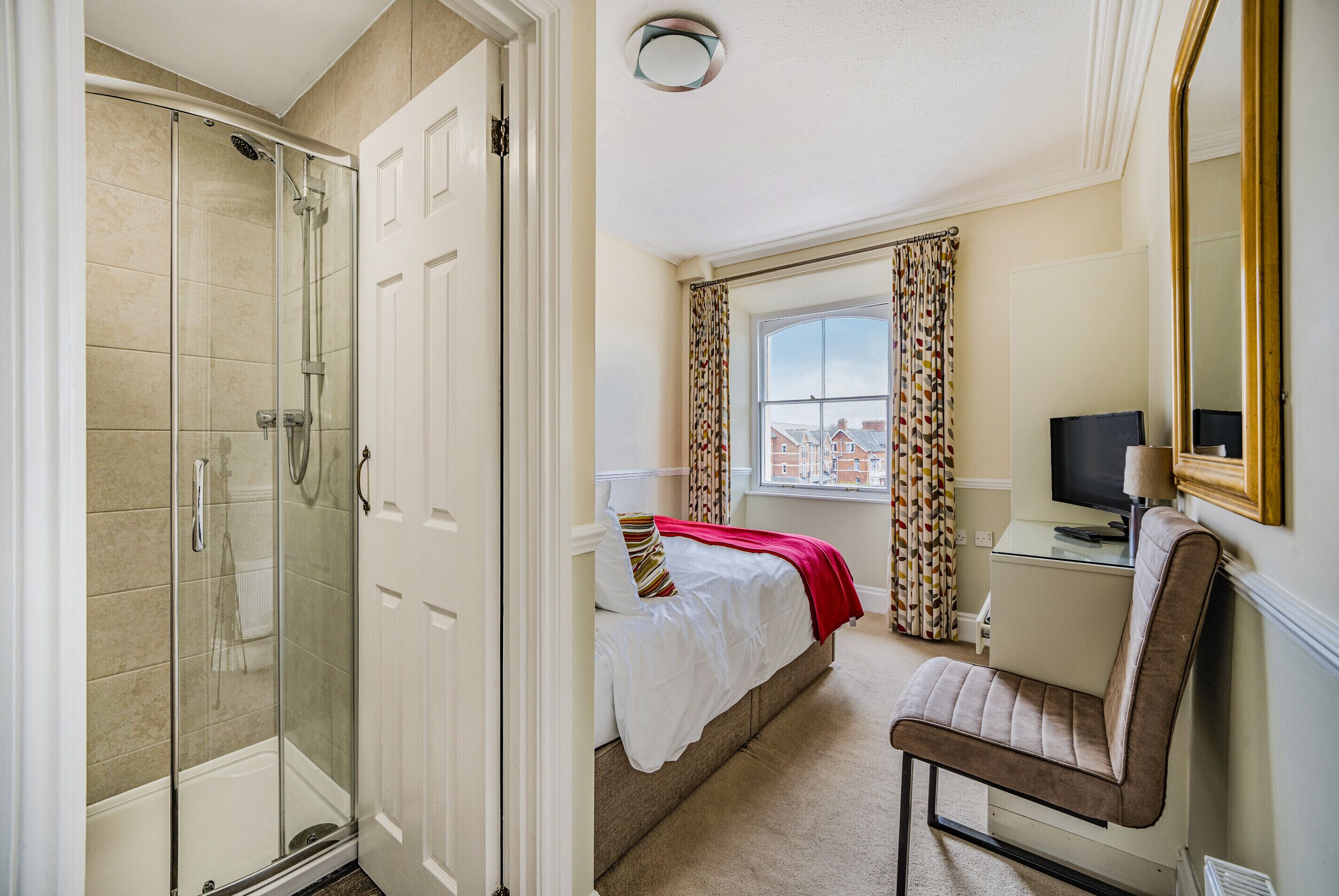 A hotel room with a glass-enclosed shower, a bed with a red blanket, a chair, a desk with a television, and a window with curtains.