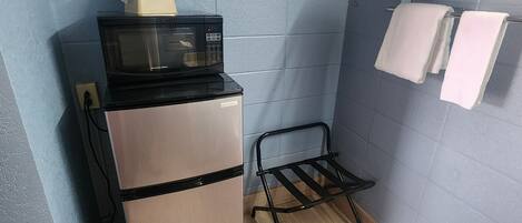 A compact kitchenette with a refrigerator, microwave, and a small shelf with a towel and a green box.