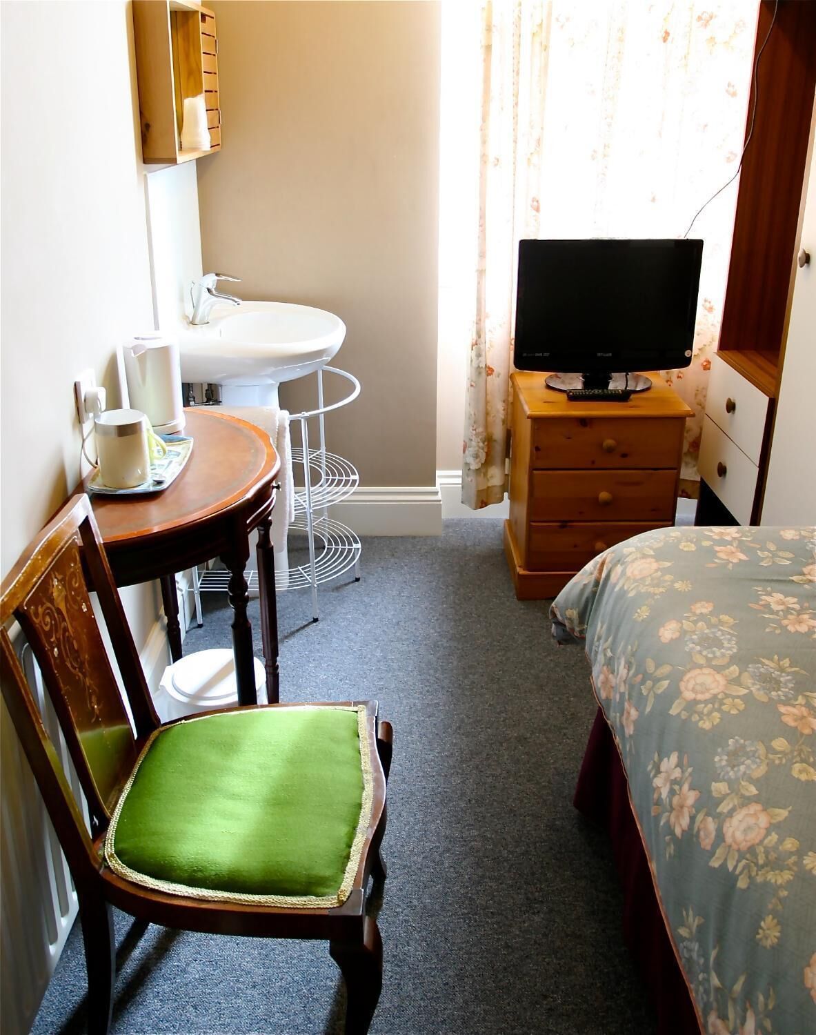 A compact hotel room with a single bed, a small round table with a chair, a television on a wooden dresser, a white sink, and a window with curtains.