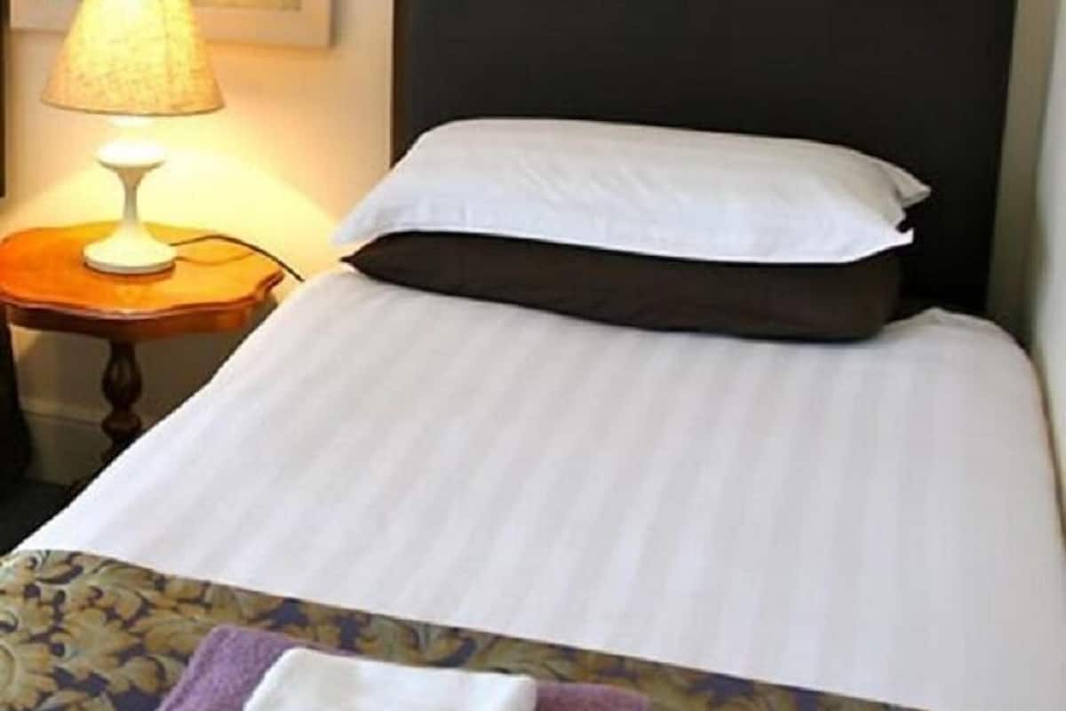 Una cama bien hecha con un cojín blanco y un cabecero oscuro, una lámpara en una mesita de noche de madera, y un cuadro en la pared.