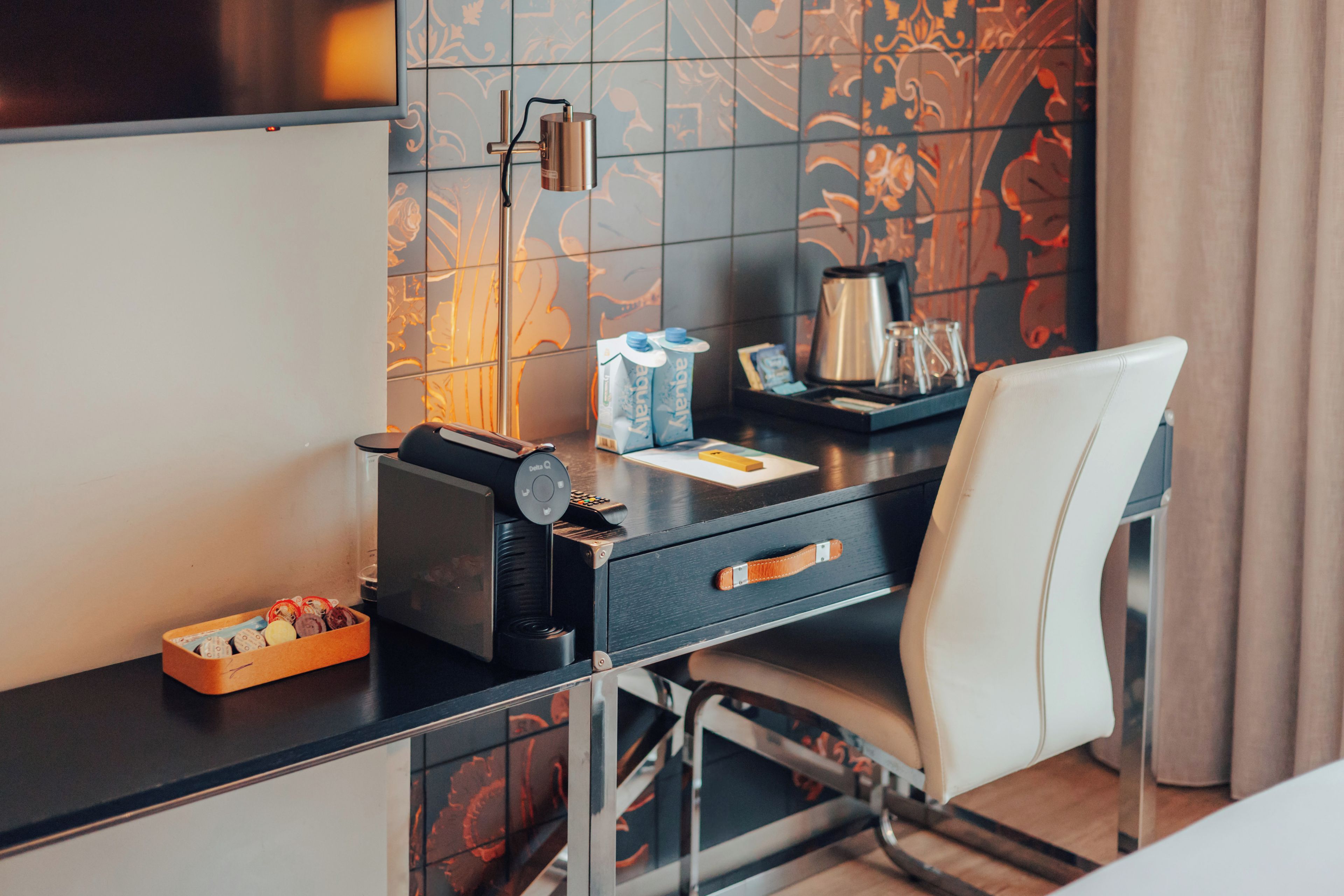 A hotel room with a desk, a chair, a coffee maker, a bottle of water, a box of tissues, and a tray with glasses and a cup.
