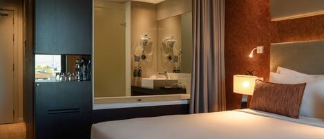 Habitación de hotel con cama, un espejo grande, mesita de noche con lámpara y vista a la ciudad por la ventana.