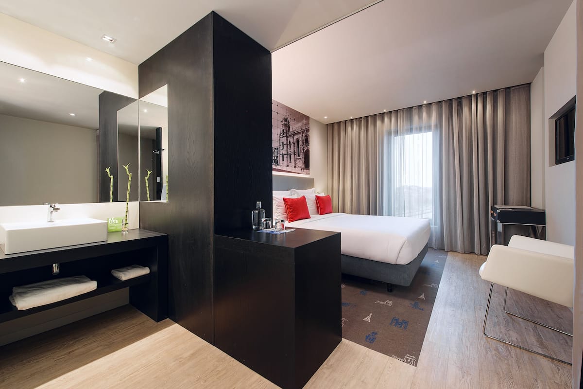 Ein modernes Hotelzimmer mit einem großen Bett, einer separaten Sitzecke, einem großen Spiegel und einem Badezimmer mit Waschbecken und Handtuchhalter.