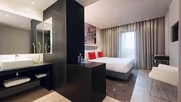 Una habitación de hotel moderna con una cama grande, una zona de estar separada, un espejo amplio y un baño con lavabo y toallero.