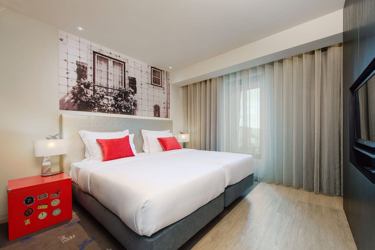 Ein modernes Hotelzimmer mit einem großen Bett, einem roten Nachttisch, einem Wandbild und einem großen Fenster mit Vorhängen.