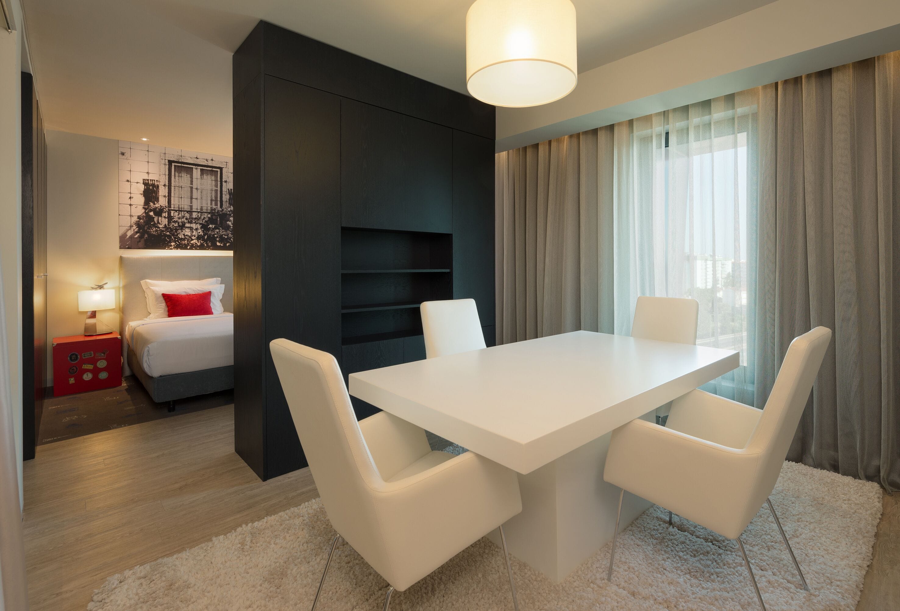 A modern hotel room with a white bed, a red accent pillow, a white dining table with four chairs, a large window with curtains, and a wall-mounted shelf.