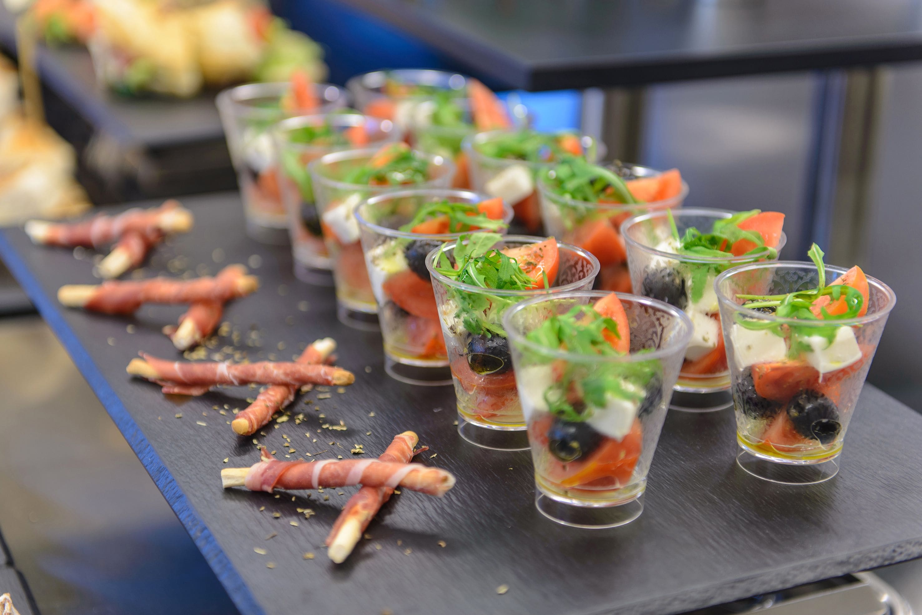 Una hilera de vasos transparentes con una ensalada y diversos ingredientes, sobre una superficie de pizarra oscura.